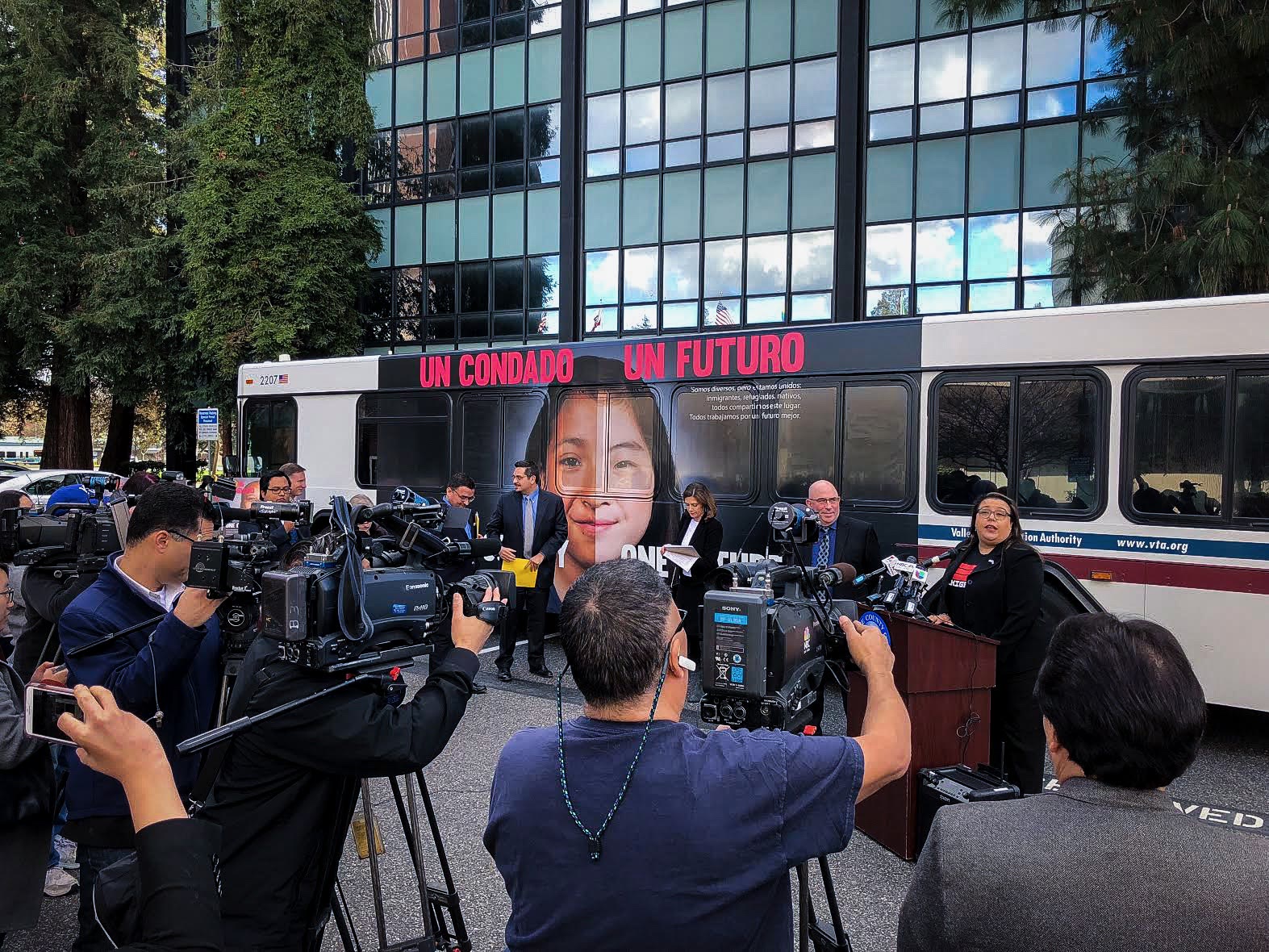 A press conference at a transit yard, a wrapped VTA bus behind the podium showing a young girl's face and the headline Un Condado Un Futuro, reporters with cameras in the foreground
